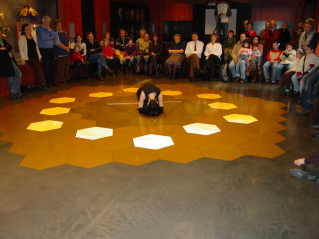 Fotografie einer Performance mit einer knienden Person in der Mitte eines Bodens aus leuchtend gelben und weißen Sechsecken. Umgeben von Publikum thematisiert das Werk „Tanz auf dem Facettenauge“ Bewegung, Raum und geometrische Strukturen.